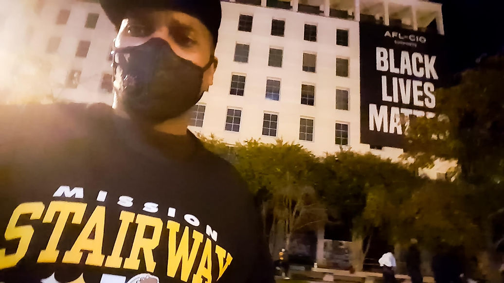 A Black man in his 30s wearing a mask and standing in front of a Black Live Matter sign in NYC.